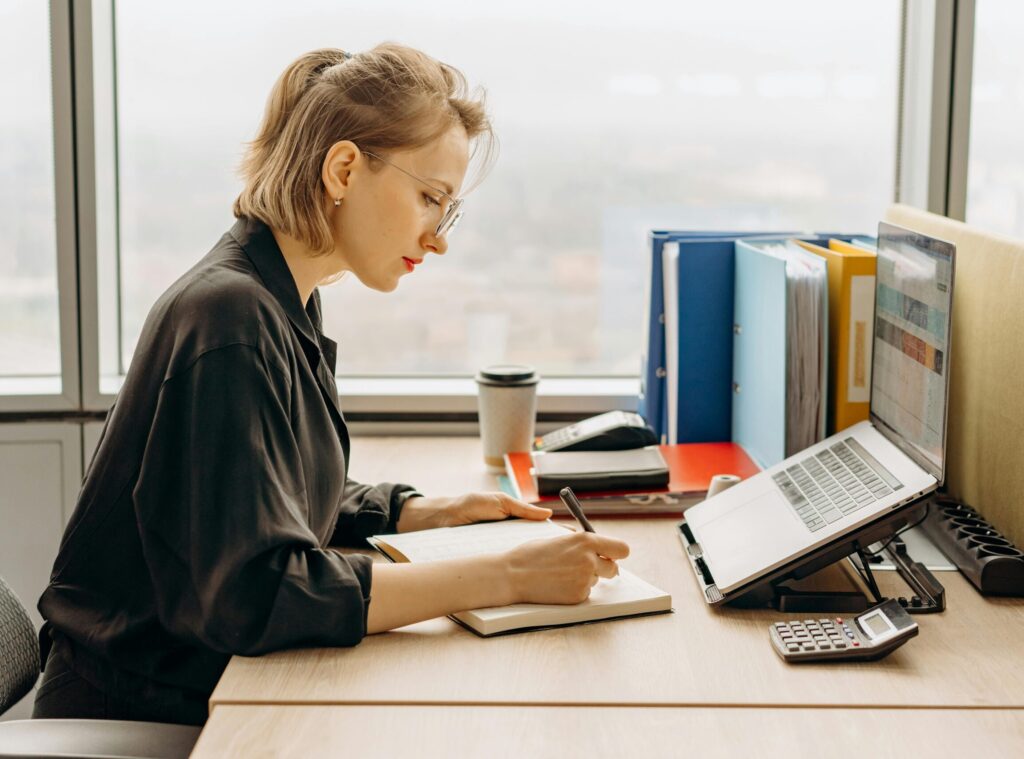 A woman sitting at a desk in an office, reviewing her bills on a laptop She is taking notes in a notebook, with office supplies, folders, a calculator, and a coffee cup on the desk. A large window in the background provides natural light.