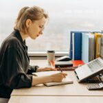 A woman sitting at a desk in an office, reviewing her bills on a laptop She is taking notes in a notebook, with office supplies, folders, a calculator, and a coffee cup on the desk. A large window in the background provides natural light.