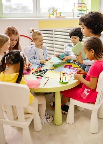 Children and teachers gathered around a table engaged in creative activities, symbolizing the importance of teaching financial literacy and foundational skills early in life.