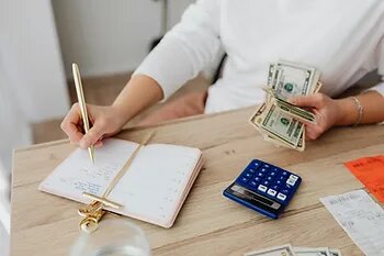 Person managing finances at a wooden desk, holding cash in one hand and writing in a notebook with the other. A calculator, receipts, and a water glass are also visible on the desk.