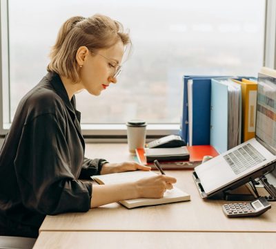 A woman sitting at a desk in an office, reviewing her bills on a laptop She is taking notes in a notebook, with office supplies, folders, a calculator, and a coffee cup on the desk. A large window in the background provides natural light.